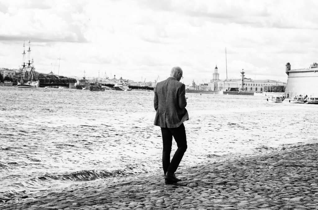 pexels-photo-8567308-8567308 A man in a blazer walks on a cobblestone path by the sea in grayscale.